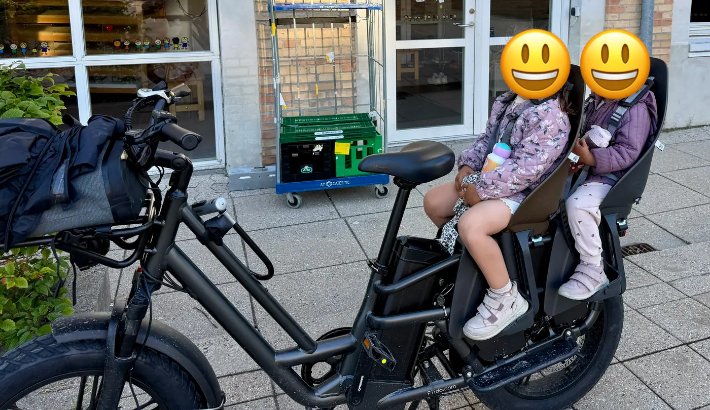 a fiido t2 longtail cargo e-bike parked on a paved forecourt outside a bakery, with two child seats mounted in line on the rear rack, both occupied by children.
